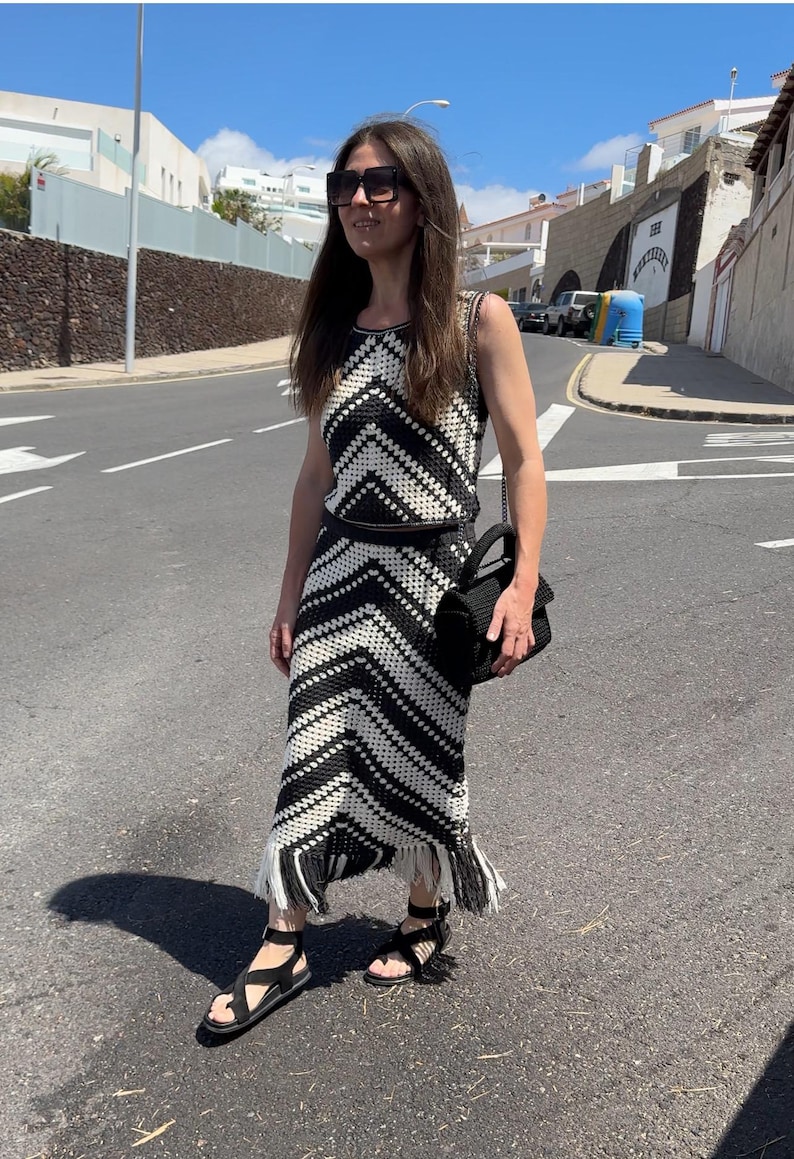 Puede incluir: Conjunto de dos piezas de crochet en blanco y negro, con un top sin mangas y una falda larga con flecos. El conjunto se combina con sandalias negras y un bolso de mano negro. El conjunto tiene un patr&oacute;n de chevr&oacute;n.