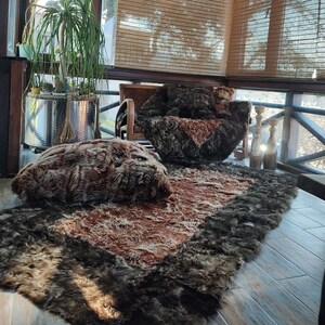 May include: A cozy indoor scene featuring a large, plush chair, a rectangular rug, and a large pillow, all with a shaggy, brown and dark grey fur-like texture. The rug and pillow are on a wooden floor, with a view of the outdoors.