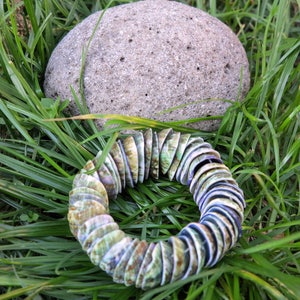 May include: A circular bracelet made of iridescent, shell-like pieces, with shades of green, blue, and tan. A gray, porous stone rests in the background, set against a backdrop of green grass.