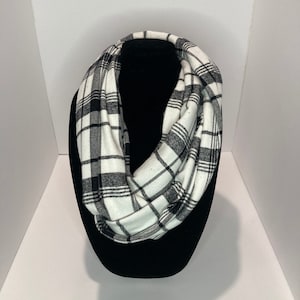 May include: A black and white plaid infinity scarf. The scarf is made of a soft fabric and is displayed on a black mannequin head. The pattern consists of black lines on a white background, creating a classic checkered design.