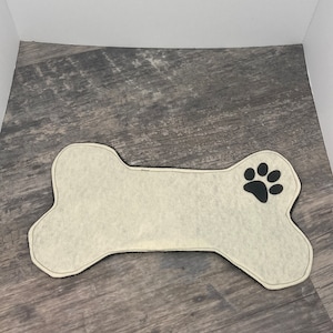 Könnte beinhalten: Eine weiße, knochenförmige Hundematte mit einem schwarzen Pfotenabdruck in der Ecke.