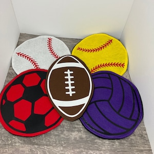 Könnte beinhalten: Fünf Filz-Sportball-Dekorationen in verschiedenen Farben und Designs. Ein weißer Baseball, ein gelber Softball, ein brauner Football, ein roter Fußball und ein violetter Volleyball.
