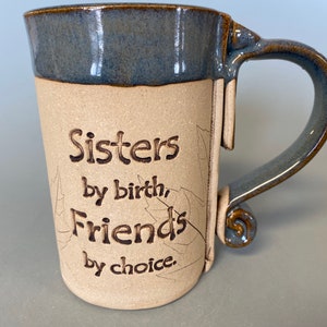 May include: A handmade ceramic mug with a blue glazed rim and handle. The mug features the text "Sisters by birth, Friends by choice" in brown lettering. The mug has a beige body with leaf-like etchings.