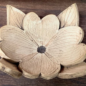 May include: A beige, carved wooden flower with a dark center and detailed petal and leaf patterns. The flower has four large petals and three leaves. The object is likely a decorative element or a piece of art.