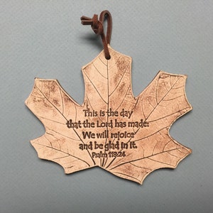 May include: A maple leaf-shaped ornament with a brown leather cord. The ornament has etched leaf veins and the text "This is the day that the Lord has made: We will rejoice and be glad in it. Psalm 118:24."