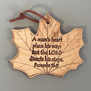 May include: A tan maple leaf-shaped ornament with a brown leather cord for hanging. The surface features dark brown engraved text that reads: A man's heart plans his way: But the LORD directs his steps. Proverbs 16:9. The leaf has visible veins.