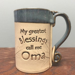 May include: A handmade ceramic mug with a blue rim and handle. The mug is beige with the words "My greatest Blessings call me Oma" etched into the side. The handle has a decorative swirl at the base.
