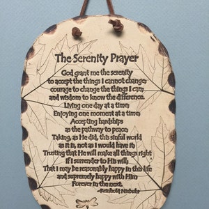 May include: A ceramic plaque with the Serenity Prayer inscribed in black lettering. The plaque has a leaf-shaped design with a brown leather cord for hanging. The text includes the author's name, Reinhold Niebuhr.
