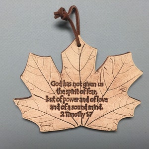 May include: A maple leaf-shaped ornament with a brown leather cord. The leaf is a light tan color with etched veins and the text "God has not given us the spirit of fear, but of power and of love and of a sound mind. 2 Timothy 1:7."