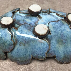 May include: A handcrafted ceramic bowl with a unique, organic shape. The bowl features a blue-green glaze with a speckled texture and is adorned with five small, round, white and brown accents. This decorative piece is ideal for serving or display.