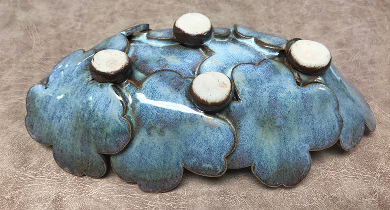 May include: A handcrafted ceramic bowl with a unique, organic shape. The bowl features a blue-green glaze with a speckled texture and is adorned with five small, round, white and brown accents. This decorative piece is ideal for serving or display.