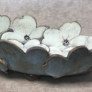 May include: A ceramic serving bowl with a floral design. The bowl is shaped like a series of white flowers with dark centers and a gray-blue rim. The bowl is resting on a brown surface.