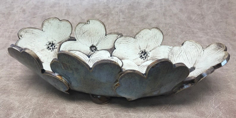 May include: A ceramic serving bowl with a floral design. The bowl is shaped like a series of white flowers with dark centers and a gray-blue rim. The bowl is resting on a brown surface.