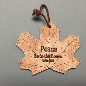 May include: A leaf-shaped ornament made of light brown leather, featuring the text "Peace be to this house. Luke 10:5." The ornament has a detailed leaf vein design and a brown leather loop for hanging.