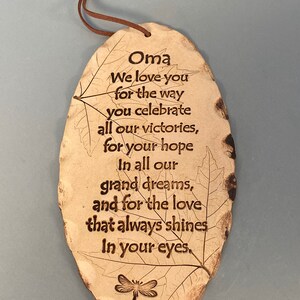 May include: A tan, leaf-shaped ceramic plaque with a brown leather cord for hanging. The plaque features the word "Oma" and an engraved message about love and appreciation, along with a dragonfly illustration.