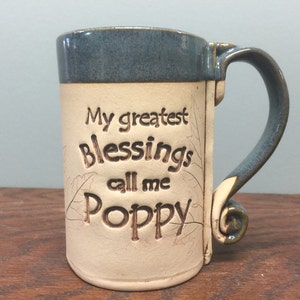May include: A handmade ceramic mug with a blue rim and handle. The mug is cream-colored with the words "My greatest Blessings call me Poppy" etched into the front. The handle has a unique, decorative swirl.