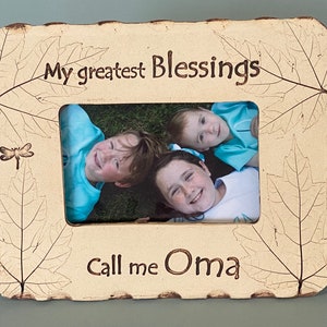 May include: A rectangular photo frame with a cream-colored, leaf-patterned border. The frame holds a photo of three smiling children. The text "My greatest Blessings" and "Call me Oma" are inscribed on the frame. A dragonfly is also visible.