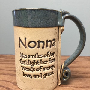 May include: A handmade ceramic mug with a blue rim and handle. The mug is beige with the word "Nonna" and a poem etched into the side. The handle has a decorative swirl at the base.