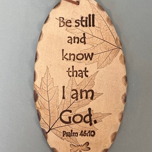 May include: Oval-shaped, light brown plaque with the engraved text "Be still and know that I am God. Psalm 46:10." Features leaf and dragonfly designs. Includes a brown leather cord for hanging. The edges have a decorative, textured border.