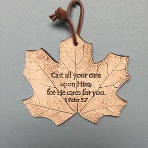 May include: A maple leaf-shaped ornament with the text "Cast all your care upon Him; for He cares for you. 1 Peter 5:7". The ornament is light brown with a leather loop for hanging.