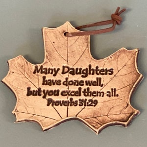 May include: A tan, leaf-shaped ornament with a dark brown leather cord. The surface features a detailed, etched leaf vein pattern and the engraved text: Many Daughters have done well, but you excel them all. Proverbs 31:29 in a dark brown font.