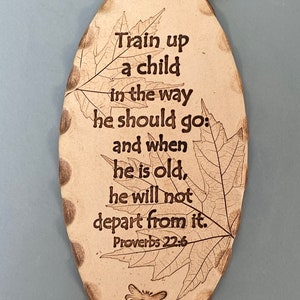 May include: Oval-shaped, beige ceramic plaque with the text "Train up a child in the way he should go: and when he is old, he will not depart from it. Proverbs 22:6." Features leaf and dragonfly designs, and a brown leather cord.