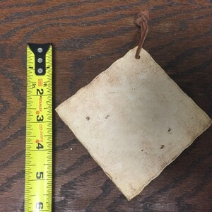 May include: A diamond-shaped, off-white tag with a brown leather string attached. The tag is approximately 3 inches wide. The tag has a distressed appearance with small dark spots. The background is a dark wood surface.