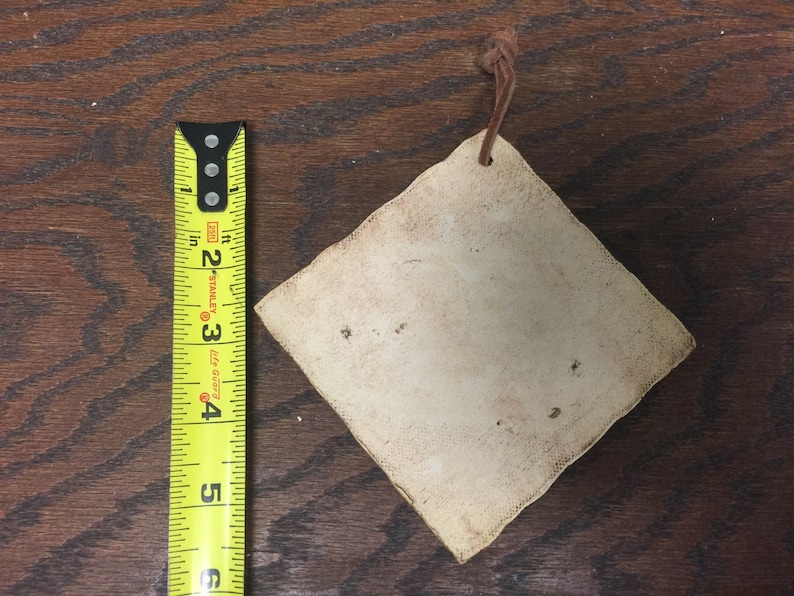 May include: A diamond-shaped, off-white tag with a brown leather string attached. The tag is approximately 3 inches wide. The tag has a distressed appearance with small dark spots. The background is a dark wood surface.