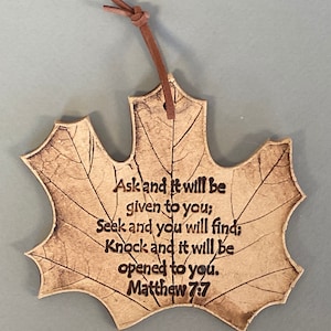 May include: A tan, maple leaf-shaped hanging ornament made of a textured material with dark brown engraved veins and the text: Ask and it will be given to you; Seek and you will find; Knock and it will be opened to you. Matthew 7:7. A brown leather cord is attached.