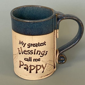 May include: A handmade ceramic mug with a blue rim and handle. The mug has a cream-colored body with the words "My greatest Blessings call me Pappy" in black script. Leaf designs are etched on the side.