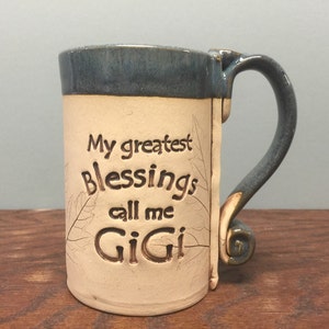 May include: Handmade ceramic mug with a cream-colored body and a blue rim and handle. The mug features the text "My greatest Blessings call me GiGi" with leaf accents. The handle has a unique, decorative design.