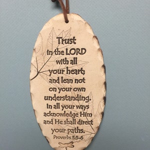May include: An oval-shaped, beige ceramic plaque with a brown leather cord for hanging. The plaque features the text "Trust in the LORD with all your heart; and lean not on your own understanding. In all your ways acknowledge Him and He shall direct your paths. Proverbs 3:5-6."