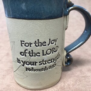 May include: A ceramic mug with a blue rim and handle, featuring the text "For the Joy of the LORD is your strength. Nehemiah 8:10." The mug has a textured, off-white surface with leaf-like designs.