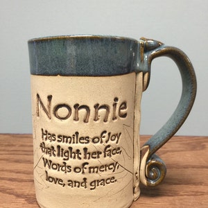 K&ouml;nnte beinhalten: Eine handgefertigte Keramiktasse mit blauem Rand und Henkel. Die Tasse ist beige mit dem Namen "Nonnie" und einem inspirierenden Zitat, das in die Oberfl&auml;che ge&auml;tzt ist. Der Griff hat ein dekoratives Blattmuster.