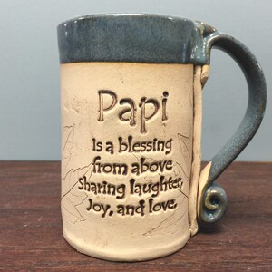 May include: A ceramic mug with a blue rim and handle, featuring the word "Papi" and the inscription "is a blessing from above sharing laughter, joy, and love." The mug has a beige body with leaf designs.