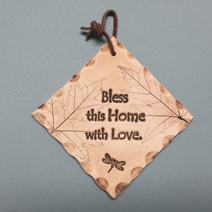 May include: A diamond-shaped, tan-colored ceramic plaque with the words "Bless this Home with Love." The plaque features leaf and dragonfly designs, and a brown leather cord for hanging.