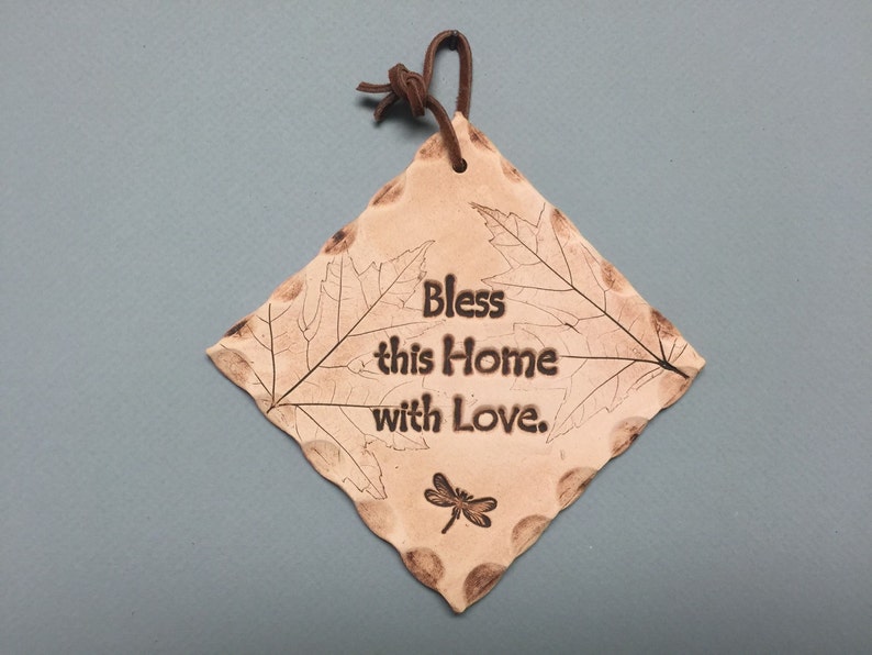 May include: A diamond-shaped, tan-colored ceramic plaque with the words "Bless this Home with Love." The plaque features leaf and dragonfly designs, and a brown leather cord for hanging.