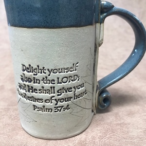 May include: A handmade ceramic mug with a blue rim and handle. The mug has a cream-colored body with the text "Delight yourself also in the LORD; and He shall give you the desires of your heart Psalm 37:4" etched on the front.