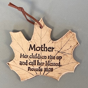 May include: A beige ceramic ornament shaped like a maple leaf with dark brown engraved text that reads Mother Her children rise up and call her blessed. Proverbs 31:28. The ornament features a detailed leaf vein pattern and a brown leather cord.