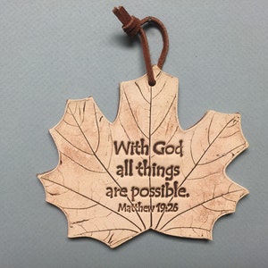 May include: A maple leaf-shaped ornament with the text "With God all things are possible. Matthew 19:26." The ornament is beige with brown detailing and a brown leather cord for hanging.