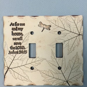 May include: Cream-colored ceramic double light switch cover with a leaf design and the text "As for me and my house, we will serve the LORD, Joshua 24:15." A dragonfly is also featured.