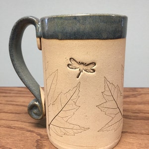 K&ouml;nnte beinhalten: Eine handgefertigte Keramik-Tasse mit hellbeigem Korpus und dunkelblauem Rand und Henkel. Die Tasse hat ein Libellen- und Blattdesign. Der Griff hat eine dekorative Spirale am Boden.