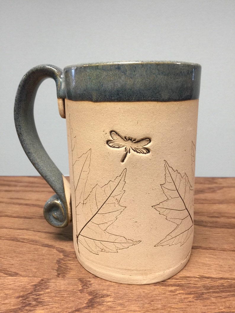 K&ouml;nnte beinhalten: Eine handgefertigte Keramik-Tasse mit hellbeigem Korpus und dunkelblauem Rand und Henkel. Die Tasse hat ein Libellen- und Blattdesign. Der Griff hat eine dekorative Spirale am Boden.