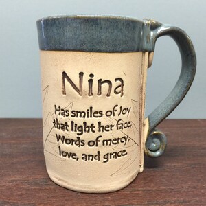 May include: A handmade ceramic mug with a blue rim and handle. The mug is beige with the name "Nina" and an inspirational quote etched into the surface. The handle has a decorative swirl.