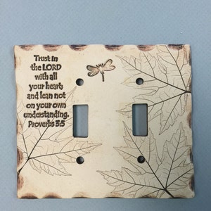 May include: Cream-colored ceramic double light switch cover with a dragonfly and leaf design. The cover has the text "Trust in the LORD with all your heart and lean not on your own understanding. Proverbs 3:5."