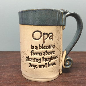 May include: A ceramic mug with a blue rim and handle, featuring the word "Opa" and the phrase "is a blessing from above, sharing laughter, joy, and love." The mug has a beige body with leaf designs.