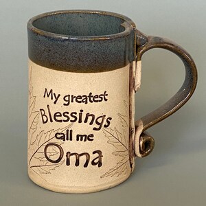 May include: Handmade ceramic mug with a blue rim and handle. The mug features the text "My greatest Blessings call me Oma" in black script, with leaf designs. The mug is beige with brown accents.