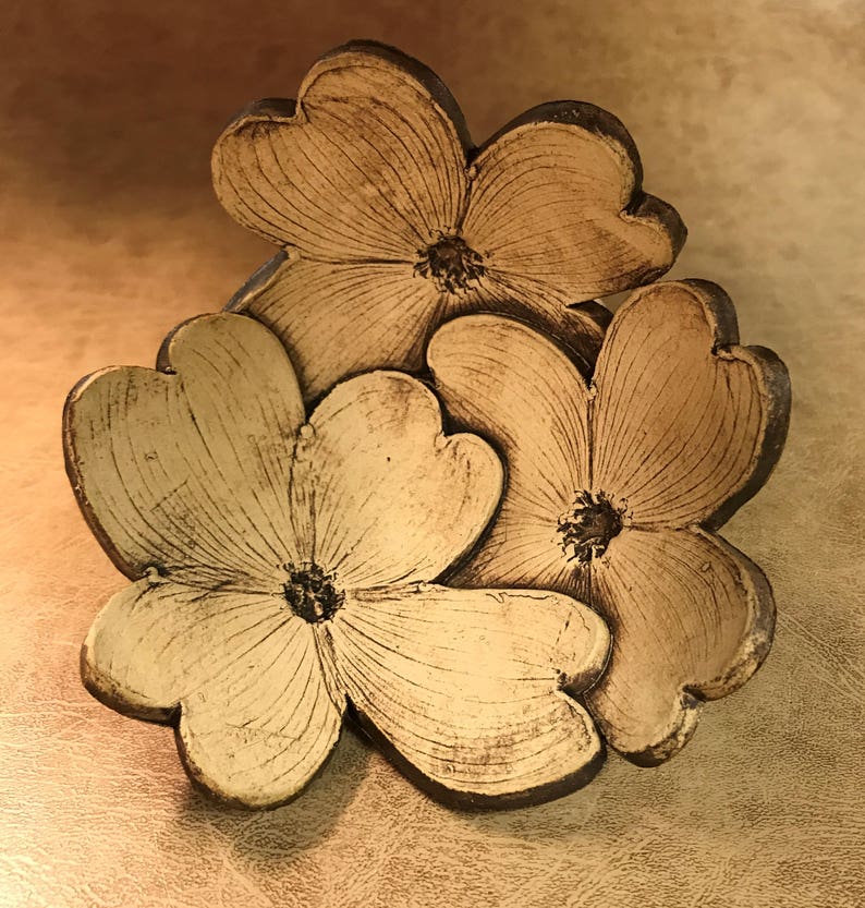 Puede incluir: Una escultura floral de cer&aacute;mica con tres flores estilizadas con p&eacute;talos en forma de coraz&oacute;n. Las flores son de color crema con contornos y centros marr&oacute;n oscuro. La escultura tiene un aspecto r&uacute;stico y hecho a mano.