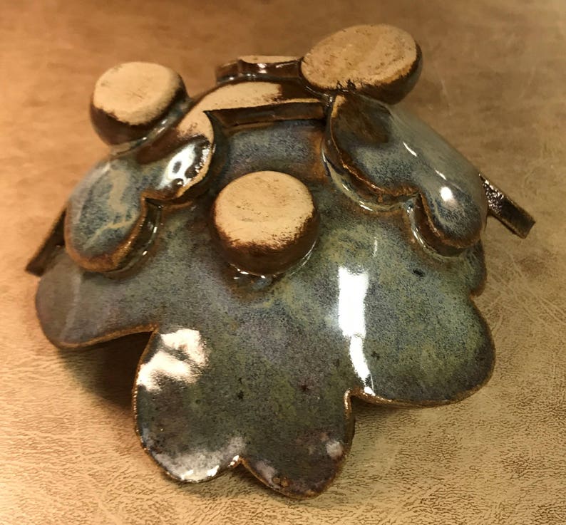 Puede incluir: Un cuenco de cer&aacute;mica hecho a mano con una forma org&aacute;nica &uacute;nica. El cuenco presenta un esmalte azul-verdoso moteado y brillante con detalles marrones. El dise&ntilde;o incluye bordes en forma de p&eacute;talo y detalles circulares en relieve.