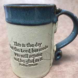 May include: A handmade ceramic mug with a blue rim and handle. The mug has a cream-colored body with the text "This is the day that the Lord has made. We will rejoice and be glad in it. Psalm 118:24."
